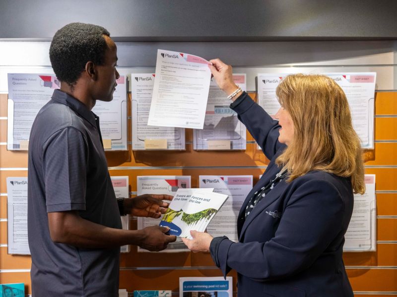 Photograph of a woman handing a man an A4 brochure from a wall of printed information.
